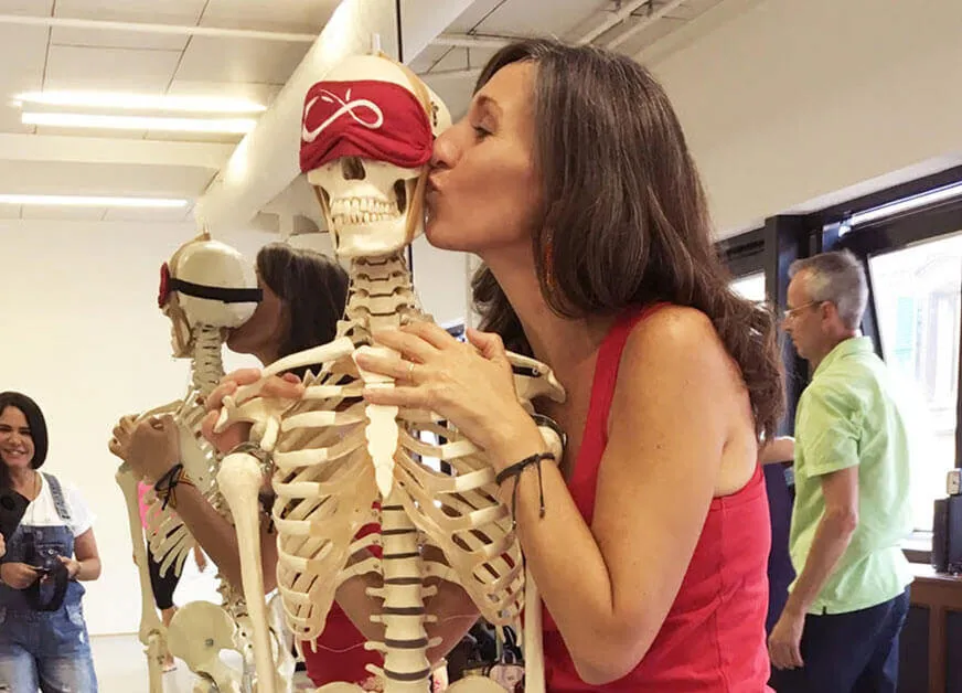 A woman in a red tank top kisses a human skeleton model.