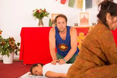 A male instructor with a headset assists a student lying face down on a mat in a yoga or bodywork class.