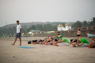 A man walking past a group of people doing a seated forward bend yoga pose on a beach.