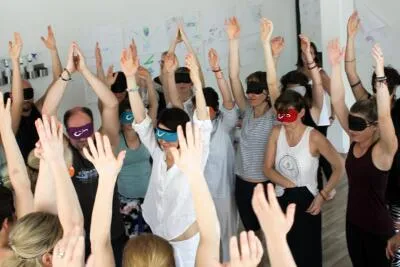 A group of people with blindfolds on and their arms raised in the air in a workshop.