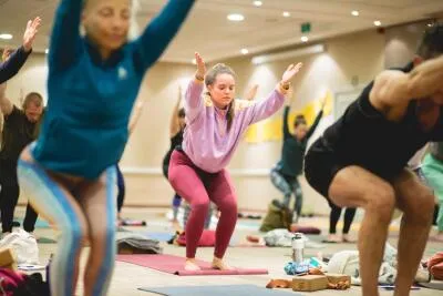 A group of people in a yoga class doing a chair pose, with a woman in a purple hoodie and red leggings as the central focus.
