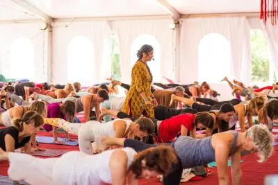 An instructor in a yellow patterned tunic walks through a large yoga class where students are holding a plank pose variation.