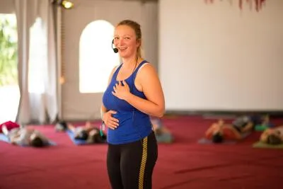 A smiling female instructor in a blue top and headset leading a yoga or fitness class.