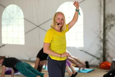 A female instructor with a headset and a yellow shirt, with her arm raised, leading a yoga or fitness class.