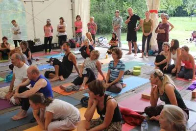 A large group of people in a yoga or meditation class, sitting and standing on mats under a tent.