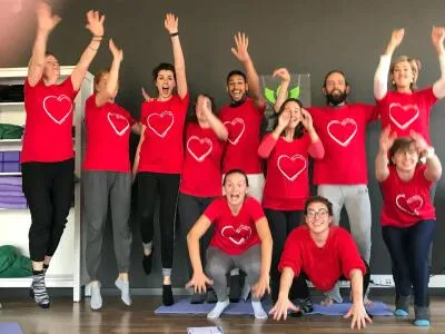 A group of people wearing red t-shirts with white hearts, smiling and posing playfully in a studio, with some jumping and others crouching.