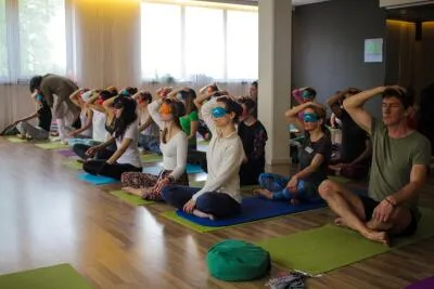 A group of people in a yoga or meditation class, many of whom are blindfolded, sitting on mats with their hands on their heads.