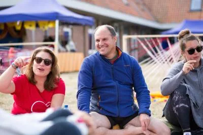 A man in a blue jacket smiles while sitting on the grass between two women at an outdoor event.