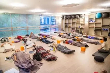A group of people lies on yoga mats, covered with blankets, in a studio during a relaxation or meditation session.