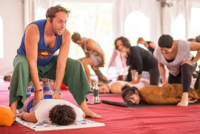 A male instructor with a headset assists a student lying face down on a mat in a yoga or bodywork class.