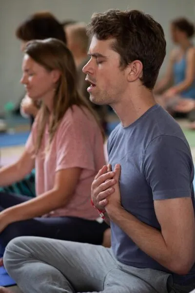 A man with his eyes closed and hands clasped over his chest, sitting in a class, possibly for yoga or meditation.