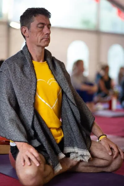 A man with his eyes closed, sitting cross-legged on the floor with a blanket or shawl draped over his shoulders.