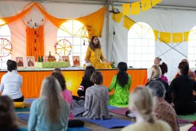 A female instructor in a yellow dress sits on a stage, speaking to a group of people sitting on yoga mats in a tent.