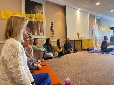 A group of people sitting on the floor on yoga mats in a workshop or class, with an instructor speaking to them.