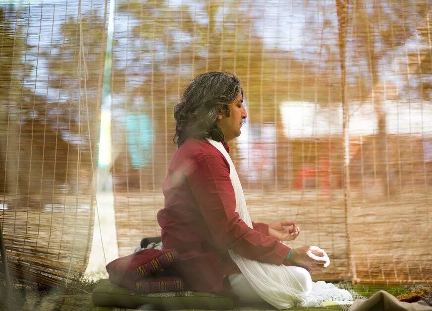 A person with long hair, wearing traditional red and white clothing, sitting in a meditative pose.