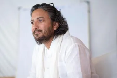 A man with a beard and long hair, wearing a white shirt and scarf, looks thoughtfully to the side.