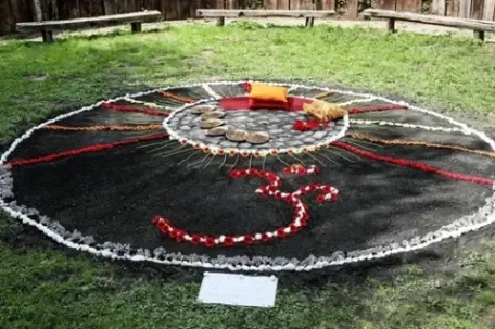 An outdoor mandala art installation made of sand, flowers, and stones, featuring the Om symbol in red petals