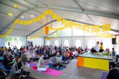 A large group of people sitting on mats in a tented room, listening to an instructor on a raised platform.
