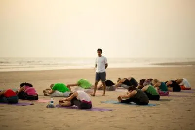 A group of people practicing yoga on a beach with a male instructor standing in the middle while the students are in a seated forward bend pose.