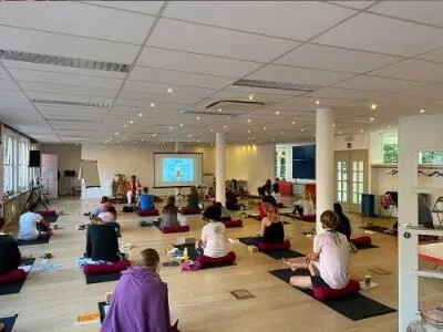 A large group of people sitting on yoga mats and cushions in a spacious room, facing a screen with a presentation.
