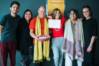 A group of six people smiling and posing with a certificate and a poster for the (Yogalife Festival 2019) in the background.