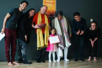 A group of adults and a young child smiling and posing for a photo with a certificate and a poster for the (Yogalife Festival 2019) in the background.