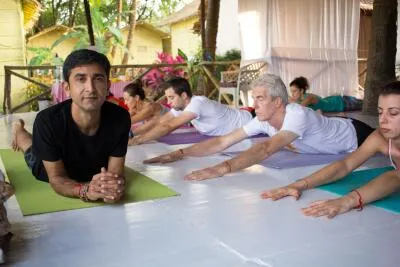 An instructor in a black shirt looks at the camera while students behind him perform a yoga pose, lying on their stomachs with their arms extended.