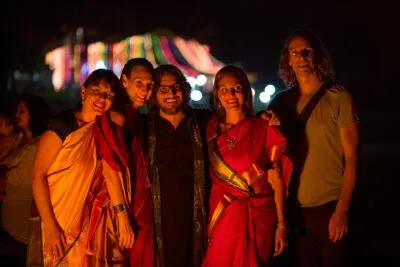 A group of five people, including two women wearing saris, smiling for a photo at night with a blurry, colorful background.