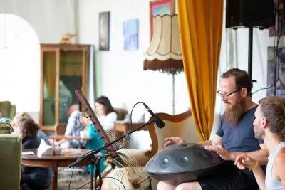 A man with a beard playing a hang drum while sitting with another man in a room.
