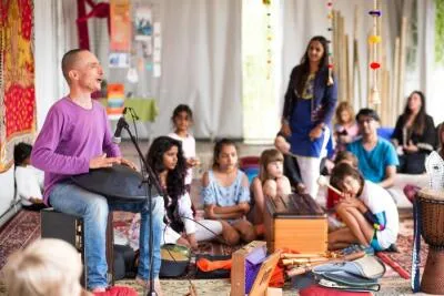 A man playing a hang drum or handpan for a group of adults and children sitting on the floor.
