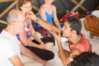 A man kneels to place a red bindi on the forehead of another man who is sitting.
