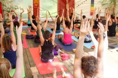 A large group of people in a yoga class with their arms raised overhead.