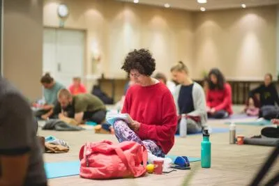A woman in a red sweater sits on the floor, writing in a notebook, with other people in the background during a class or workshop.
