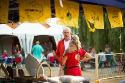 An older man in an orange jacket talking to a woman with a long braid at an outdoor festival.