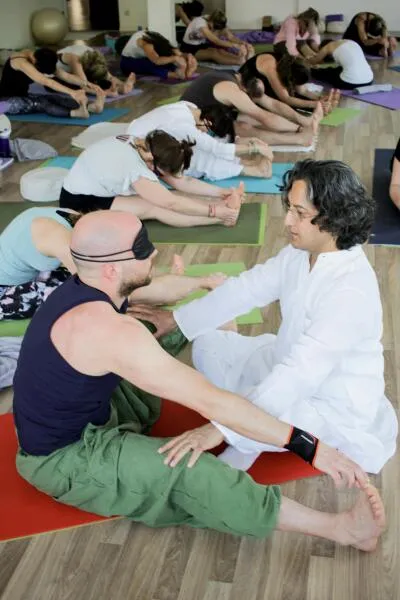 A man with a blindfold on is guided by an instructor in a yoga class while doing a seated forward fold.