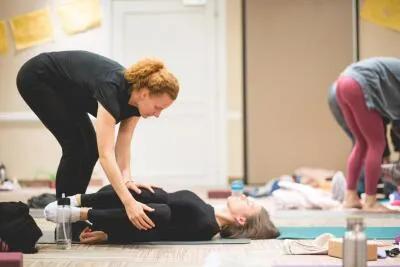 A yoga instructor assists a student lying on a mat in a knees-to-chest pose.