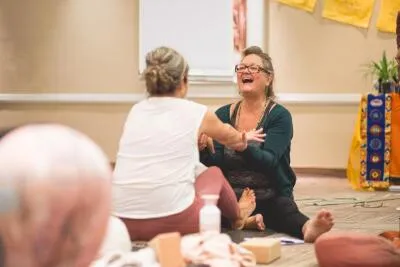 A woman with glasses and long necklaces laughing while sitting on the floor with another woman in a yoga or meditation class.