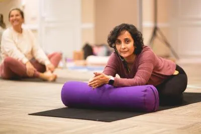 A woman in a yoga class kneeling on a mat, resting her forearms on a purple bolster, looking towards the camera.