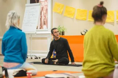 A male instructor sits cross-legged on a yoga mat, speaking to two students in a classroom setting.