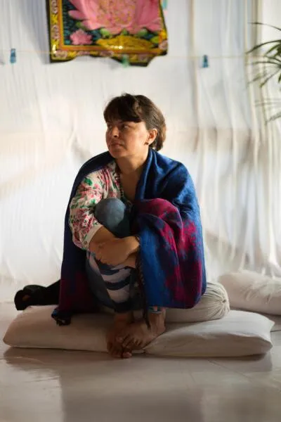 A woman sitting on the floor wrapped in a blue and red blanket, with her knees pulled up to her chest.