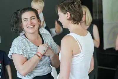 A woman laughing while carrying a baby on her back, talking to another woman in a white tank top.