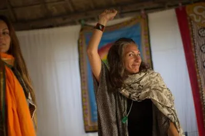 A smiling woman with her hair tied up and one arm raised, wearing a scarf and jewelry, with another woman partially visible on the left.