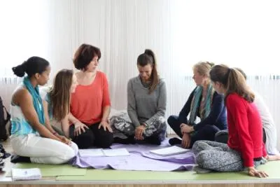 A group of women sitting in a circle on the floor, possibly in a class or meeting, with papers in front of them.