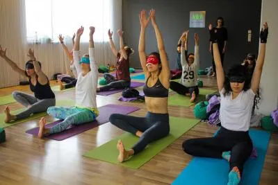 A group of people in a yoga class doing a seated pose with their arms raised, while wearing blindfolds.