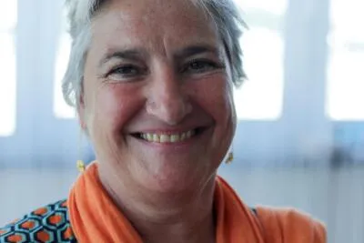 A close-up of a smiling woman with gray hair, wearing an orange scarf and long earrings.
