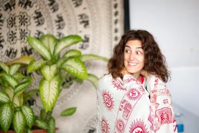 A woman with curly hair, wrapped in a white blanket with a red pattern, smiling and looking over her shoulder.