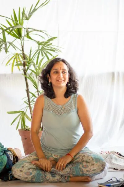 A woman with a red bindi on her forehead sits cross-legged on the floor, smiling slightly, with a potted plant behind her.