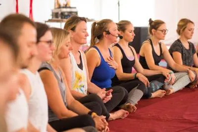A group of people sitting on the floor in a row, with one woman in the middle wearing a microphone headset.