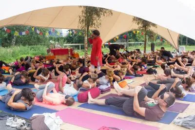 A large group of people lying on yoga mats under a tent, doing a knees-to-chest pose, with an instructor standing nearby.