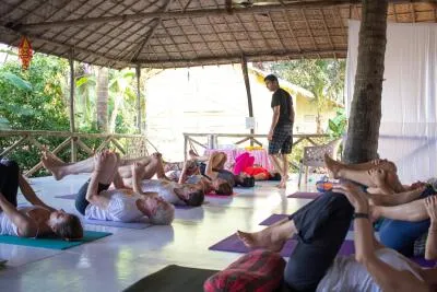 A male instructor walking among students lying on their backs on yoga mats in a knees-to-chest pose in a thatched-roof studio.
